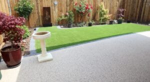 A backyard with artificial grass, surrounded by potted plants and colorful flowers along a wooden fence. A bird bath stands on a speckled patio.