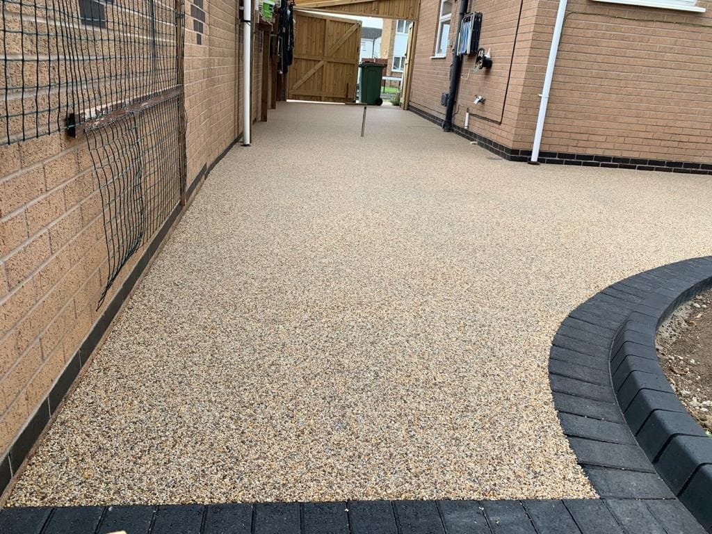 A clean, beige gravel driveway leads through a narrow passage between brick walls. A curved, dark brick border edges the right side, adding contrast.
