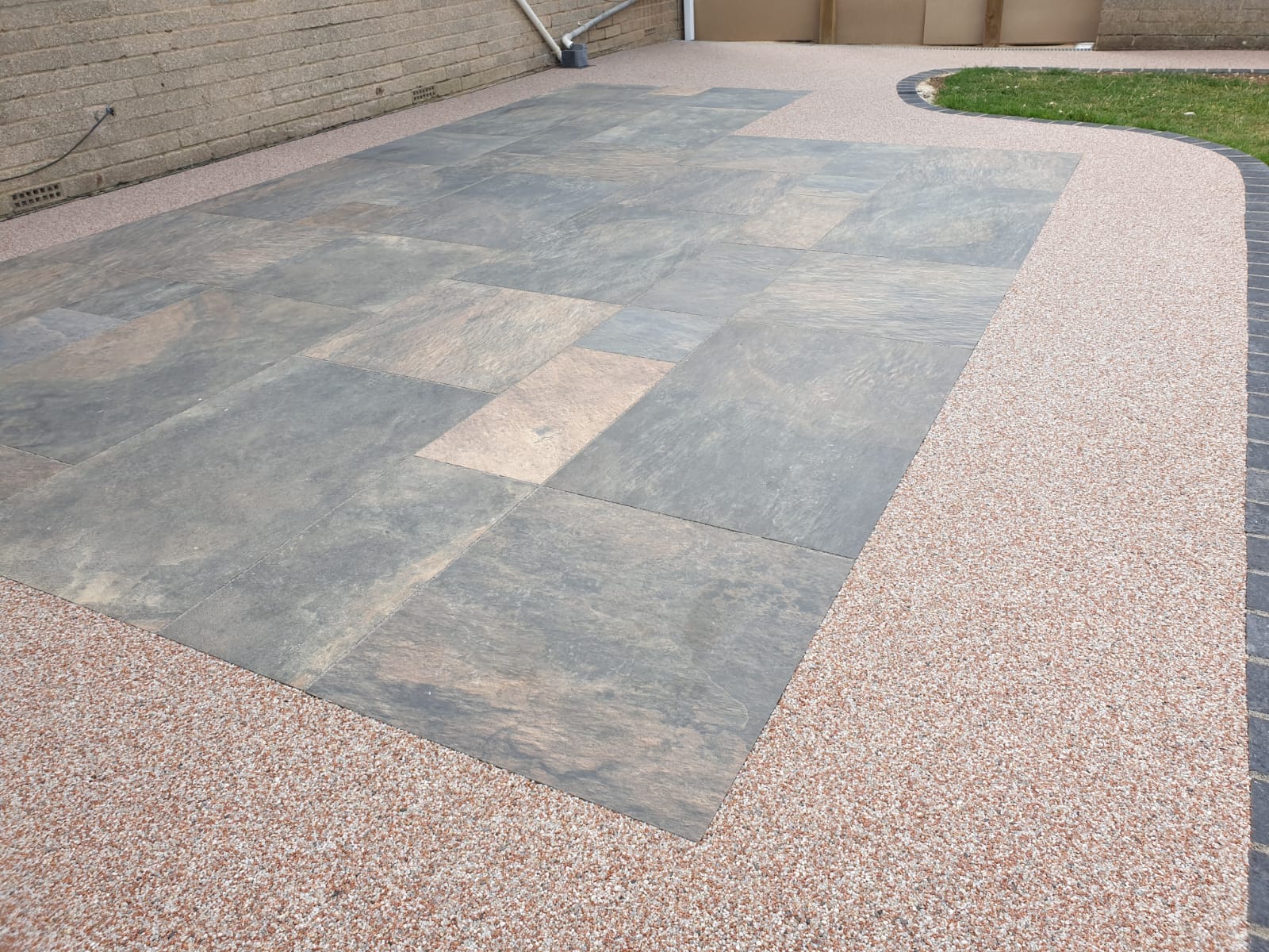 A paved courtyard with large, textured stone tiles in the center, surrounded by small pebbled concrete. Grass borders one side, creating an orderly, calm scene