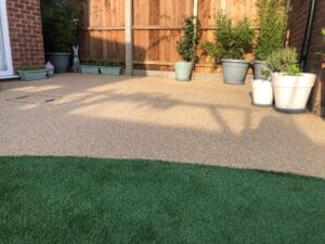 A sunlit patio with beige textured flooring, bordered by a wooden fence. Potted plants line the perimeter, and artificial grass covers the foreground.
