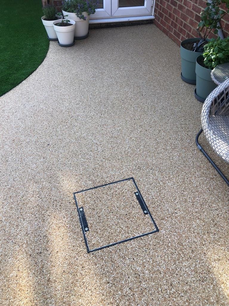 A textured beige patio with a square access panel is in the foreground. Potted plants sit near a white door and on a red brick wall, creating a calm atmosphere
