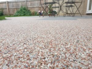 Close-up of a textured pebble patio under a cloudy sky, with blurred black metal chairs and a wooden fence in the background, creating a tranquil outdoor setting.
