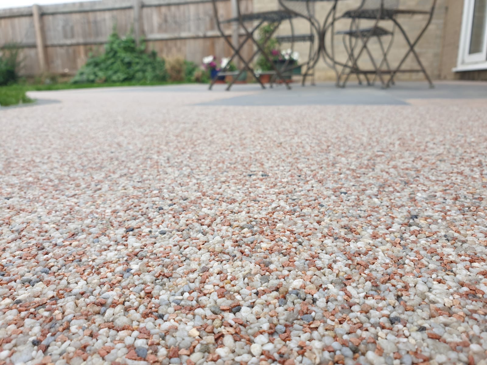 Close-up of a textured pebble patio under a cloudy sky, with blurred black metal chairs and a wooden fence in the background, creating a tranquil outdoor setting.