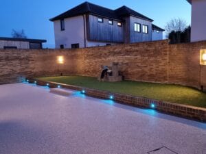 Modern backyard at dusk with a curved brick wall, lit water feature, artificial grass, and ambient blue lights along the walkway, creating a serene atmosphere.