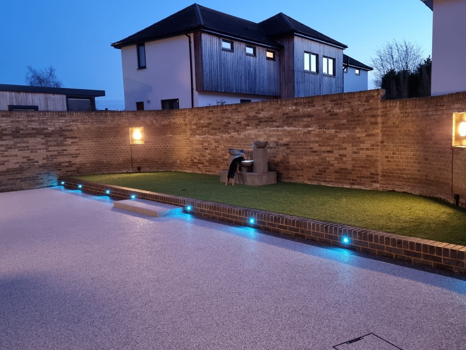 Modern backyard at dusk with a curved brick wall, lit water feature, artificial grass, and ambient blue lights along the walkway, creating a serene atmosphere.
