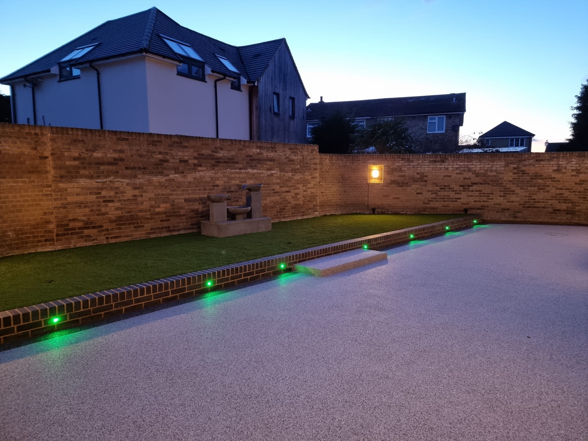 Modern backyard with a gravel area, brick walls, and subtle green lights lining the path. Evening sky creates a calm atmosphere